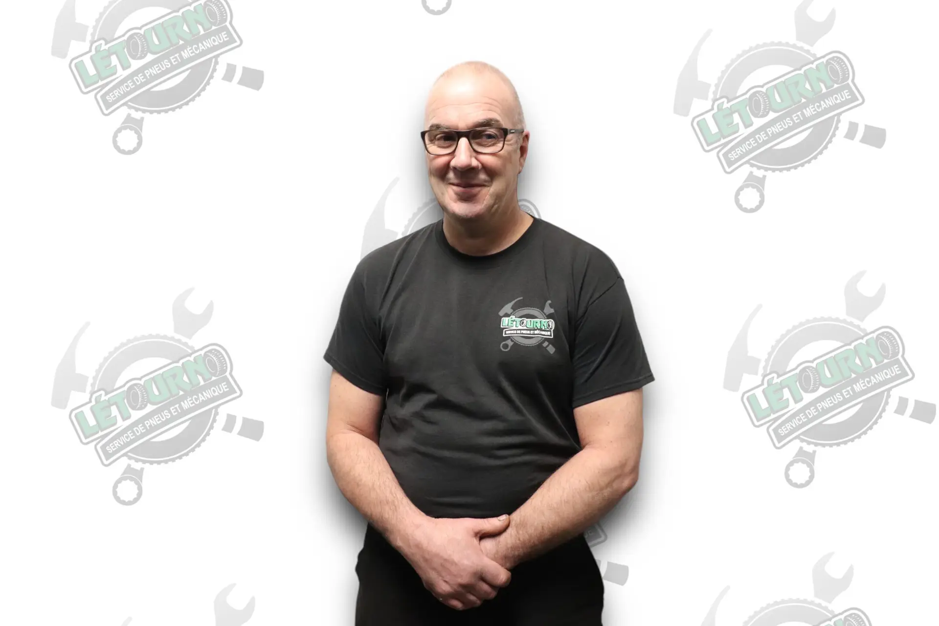 Un homme portant des lunettes et un t-shirt noir se tient debout, souriant, sur un fond blanc orné de plusieurs logos « Le TournO » défraîchis représentant des clés et des engrenages.