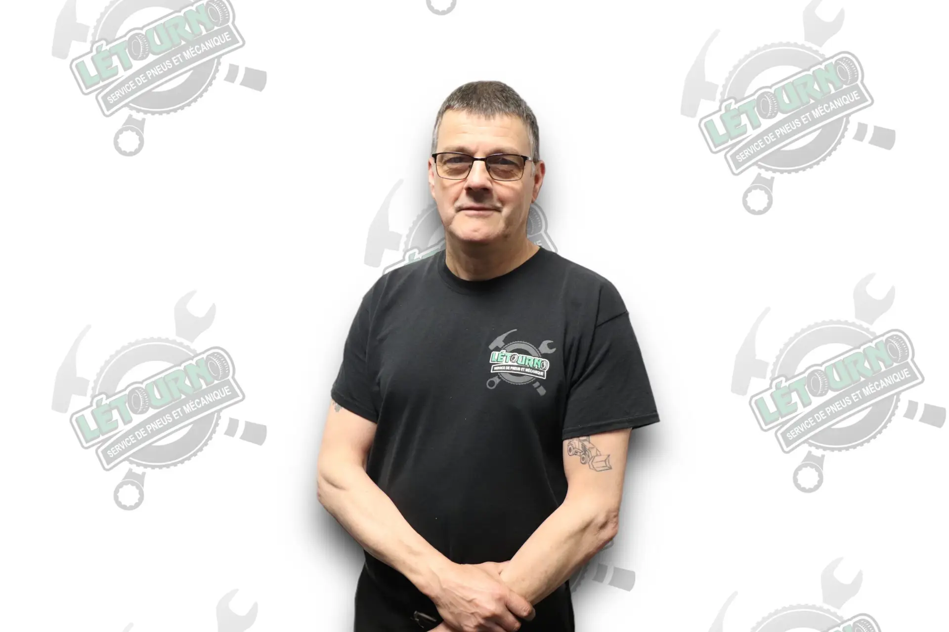 Une personne portant des lunettes et un t-shirt noir « Le Fourno » se tient debout devant un fond blanc orné de logos « Le Fourno » répétés, le regard tourné vers l'appareil photo, les mains jointes devant elle.