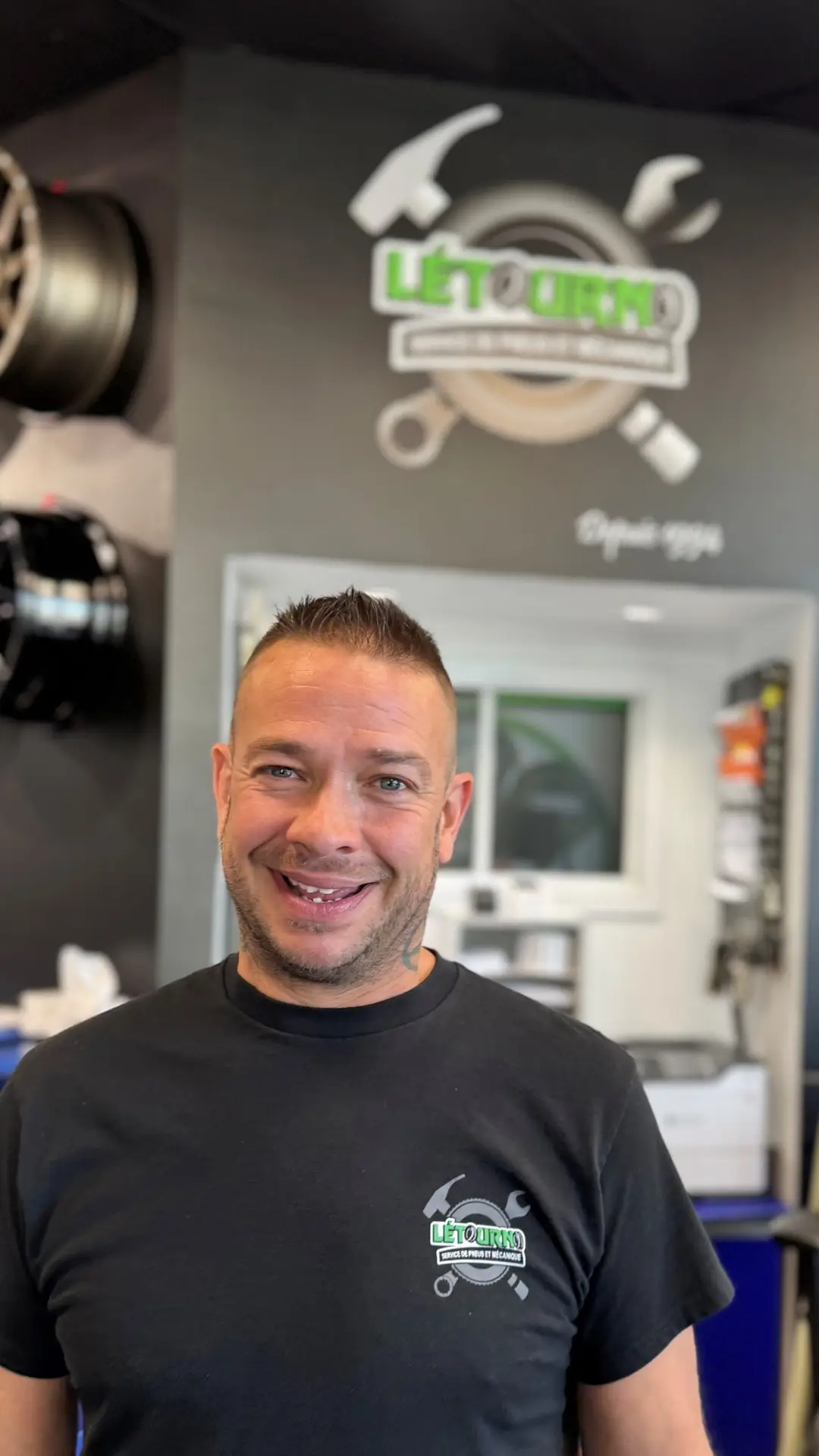 Un homme souriant, vêtu d'une chemise noire, se tient debout dans un garage automobile ; derrière lui, sur le mur, on aperçoit une enseigne « Lift Turn » ainsi qu'un logo représentant des outils de mécanicien. À l'arrière-plan, on distingue des jantes de voiture et un comptoir d'accueil.