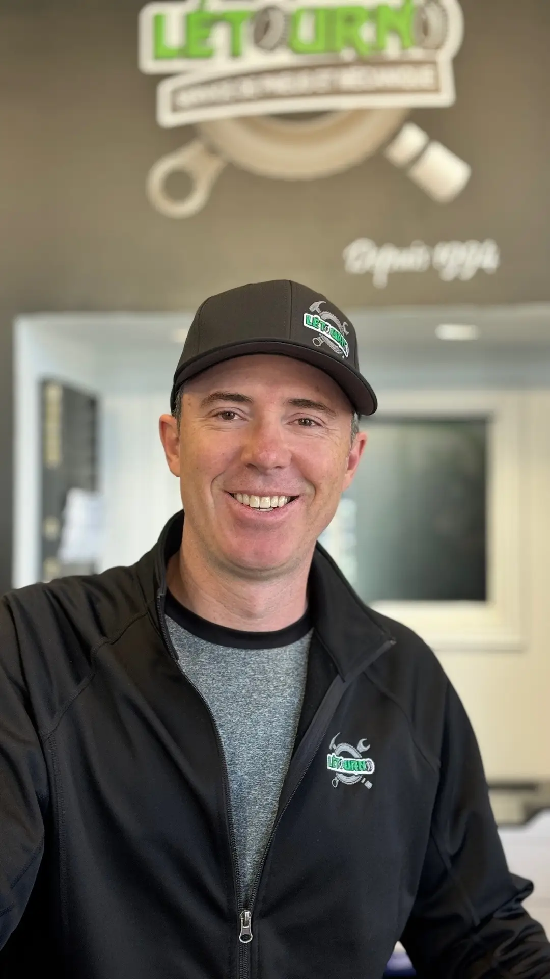 Un homme souriant, vêtu d'une veste noire et d'une casquette de baseball assortie arborant un logo vert et blanc, se tient à l'intérieur, avec une enseigne floue en arrière-plan.