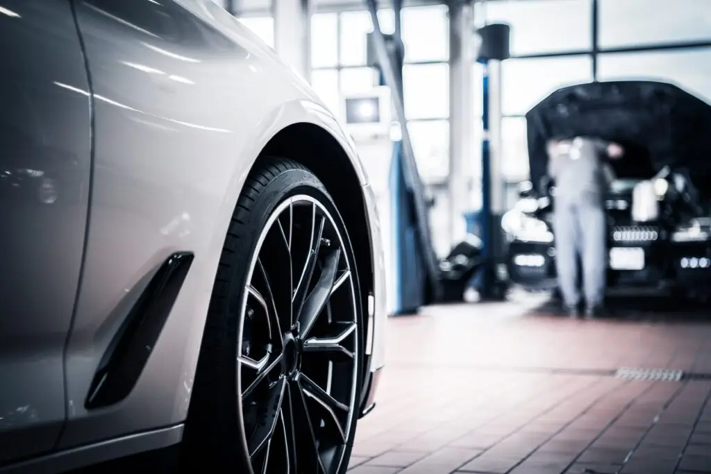 Gros plan sur une roue et un panneau latéral d'une voiture dans un garage moderne et bien éclairé, avec un mécanicien travaillant sur un autre véhicule à l'arrière-plan, flou.