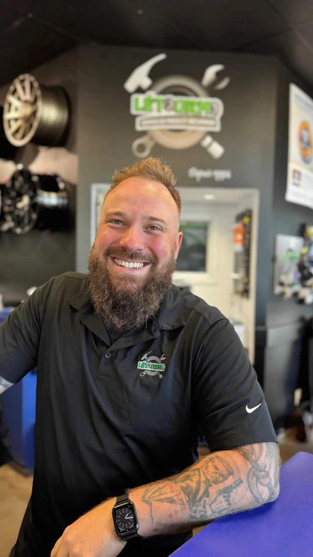 Un homme souriant, barbu, vêtu d'un polo noir arborant le logo « Lift King », se tient debout dans un garage automobile. On aperçoit à l'arrière-plan des roues de voiture et du matériel, ainsi qu'une enseigne portant l'inscription « Lift King ».