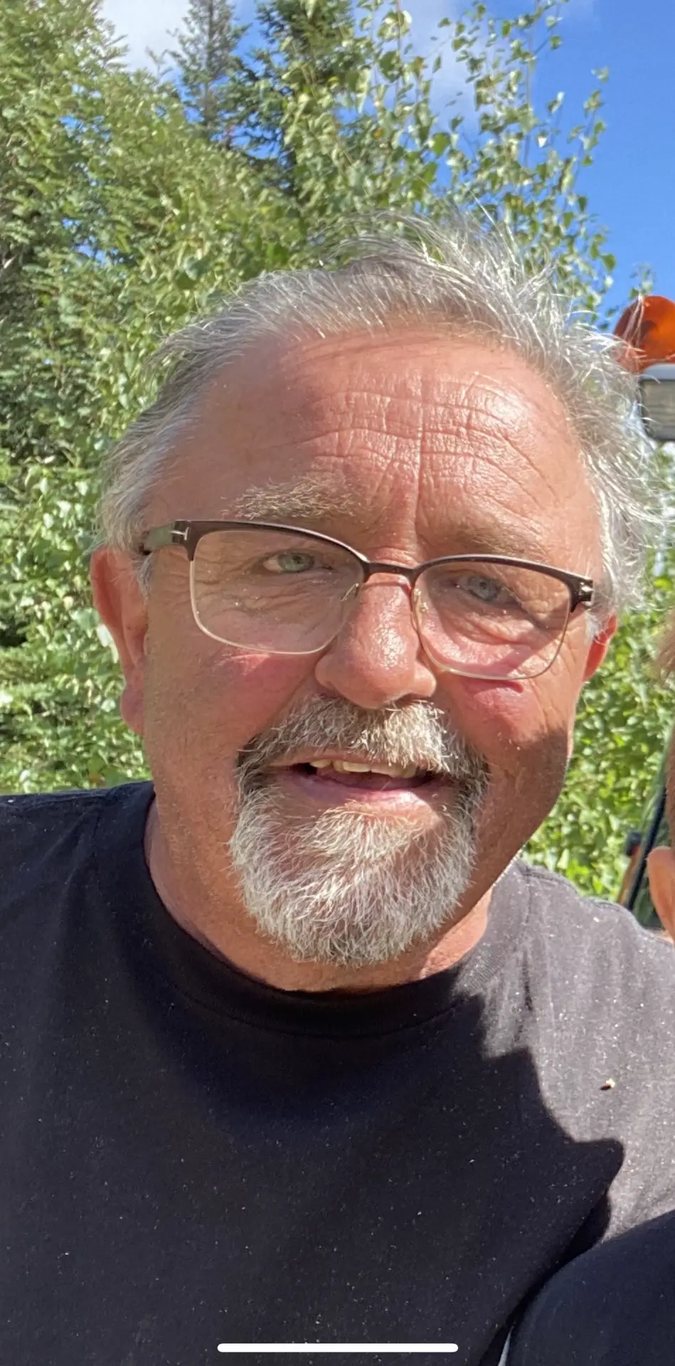 Un homme aux cheveux courts gris, portant des lunettes et une barbichette grise, sourit à l'extérieur. Il porte une chemise noire et se tient debout devant des arbres verts, sous un ciel bleu.