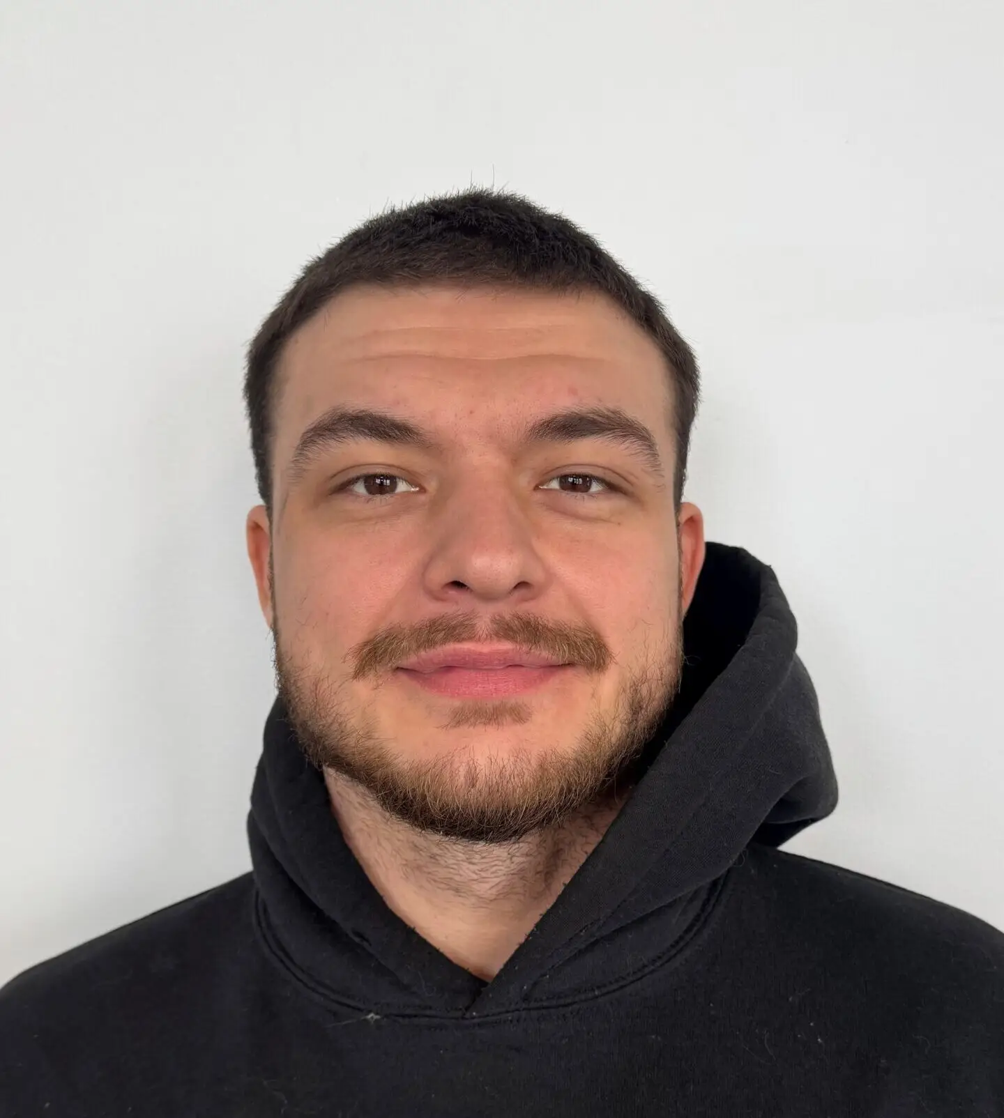 Un jeune homme aux cheveux courts et foncés, portant une barbe, est vêtu d'un sweat à capuche noir et se tient debout devant un fond blanc uni, regardant directement l'appareil photo avec une expression neutre.