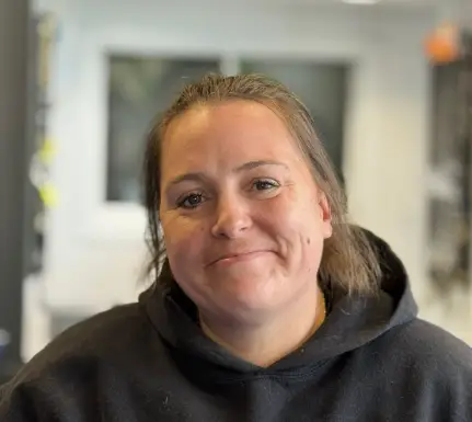 Une femme aux cheveux châtain clair attachés en arrière, vêtue d'un sweat à capuche noir, sourit doucement à la caméra dans un intérieur très éclairé.
