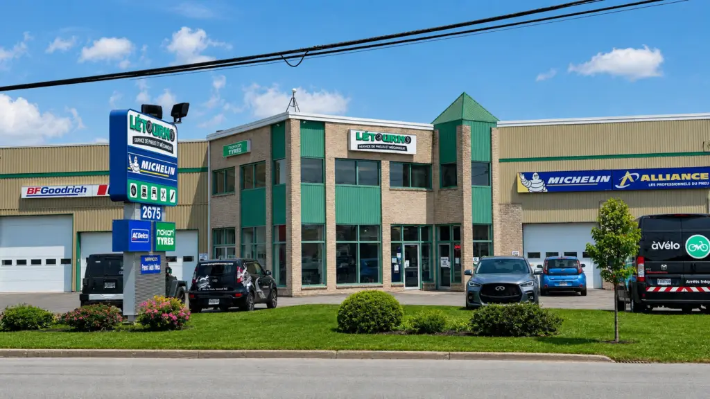 Un centre de service automobile moderne, doté de grandes baies vitrées, d'un toit végétalisé et d'enseignes pour les pneus Michelin, BF Goodrich et Alliance ; plusieurs voitures sont garées devant, par une journée ensoleillée.