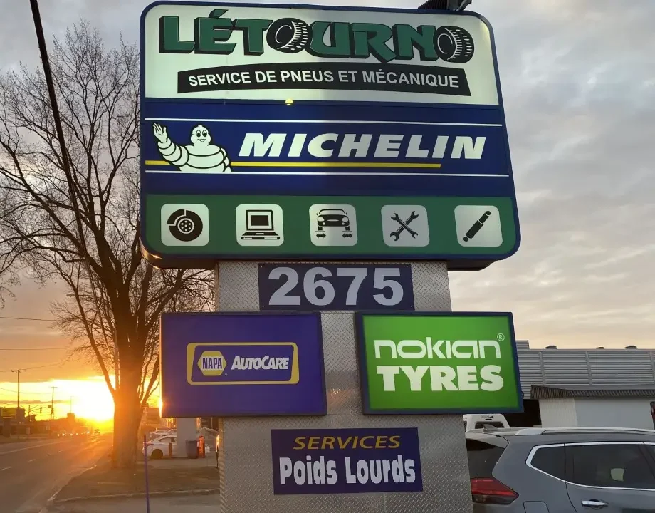 Un panneau de signalisation indiquant « Létourno Service de Pneus et Mécanique », avec les logos Michelin, Nokian Tyres et NAPA AutoCare. Le panneau mentionne les services automobiles proposés ainsi que l'adresse « 2675 ». Le soleil se couche en arrière-plan.